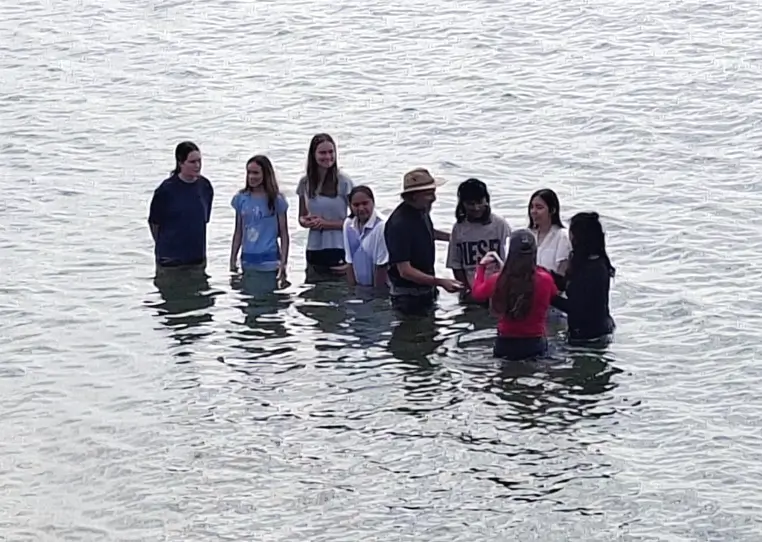 A group of young people standing in the water at a beach, preparing to be baptised